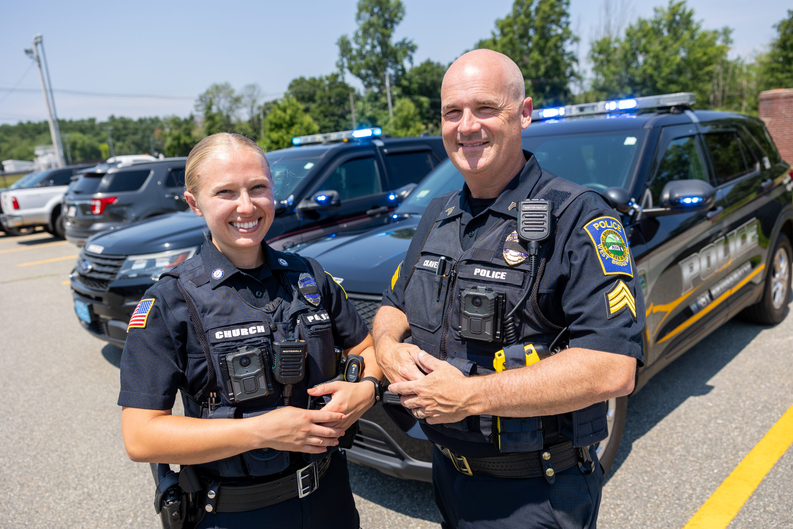 A male and female officer stand in front of two police cruisers.